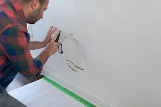 Man preparing to repair the wall. He is cutting a square hole in the wall to replace the damaged sheetrock